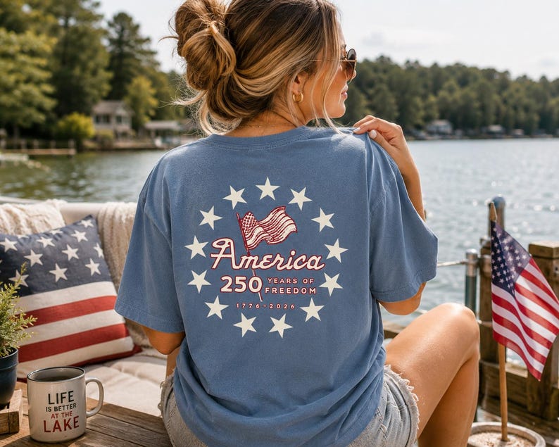 May include: A blue short-sleeve t-shirt featuring a graphic on the back with a circle of white stars surrounding a waving American flag, the text America 250 Years of Freedom, and the dates 1776-2026 printed in white and red ink.