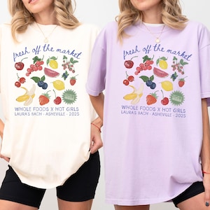 May include: Two oversized t-shirts, one cream and one lavender, featuring a fruit-themed graphic and the text "fresh off the market." The design includes cherries, berries, lemons, watermelon, and bananas. The text "WHOLE FOODS X HOT GIRLS LAURA'S BACH - ASHEVILLE - 2025" is also printed.