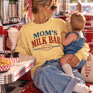 May include: A pale yellow long-sleeve sweatshirt featuring the text MOM'S MILK BAR and OPEN 24/7 in blue and red lettering. The garment is paired with blue denim jeans and worn with a red and white checkered hair ribbon and white sneakers.
