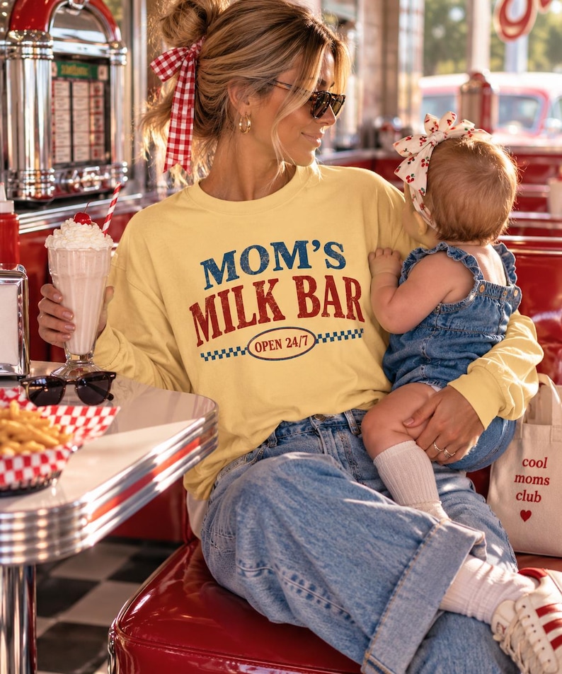 May include: A pale yellow long-sleeve sweatshirt featuring the text MOM'S MILK BAR and OPEN 24/7 in blue and red lettering. The garment is paired with blue denim jeans and worn with a red and white checkered hair ribbon and white sneakers.