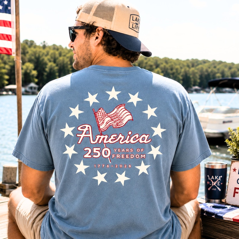 May include: A dusty blue short-sleeve t-shirt featuring a back graphic of a circle of white stars surrounding a waving American flag. The text reads America 250 Years of Freedom 1776-2026 in red and white lettering. The shirt is worn with a tan and navy cap.