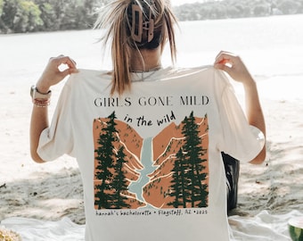 May include: Cream-colored t-shirt with the text "GIRLS GONE MILD in the wild" and a graphic of a mountain scene with trees and a waterfall. The text "hannah's bachelorette • Flagstaff, AZ • 2025" is also printed on the shirt.
