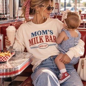 May include: A cream-colored long-sleeve sweatshirt featuring the blue and red text "MOM'S MILK BAR" and "OPEN 24/7" inside an oval with a blue and white checkered border. The sweatshirt has a crew neckline and a relaxed fit design.