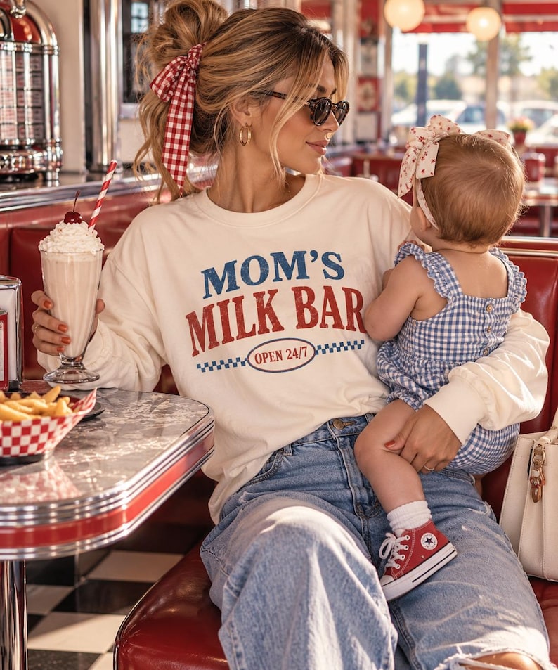 May include: A cream-colored long-sleeve sweatshirt featuring the blue and red text "MOM'S MILK BAR" and "OPEN 24/7" inside an oval with a blue and white checkered border. The sweatshirt has a crew neckline and a relaxed fit design.