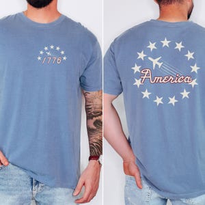 May include: A blue t-shirt with a white star design and the text "1776" on the front. The back of the shirt has a white star design with a red airplane and the text "America".