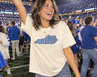 May include: A cream-colored short-sleeve t-shirt featuring a blue and white gingham patterned graphic of the state of Kentucky centered on the chest. The shirt has a crew neckline and a relaxed fit, worn with blue denim jeans.