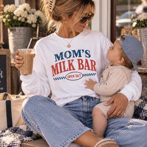 May include: A white long-sleeve crewneck sweatshirt featuring the text "MOM'S MILK BAR" in blue and red distressed lettering, with "OPEN 24/7" inside an oval graphic below. The design includes a small blue and white checkered pattern accent.