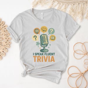 May include: A light gray t-shirt with a retro microphone graphic and the text "I speak fluent trivia". The graphic features a microphone with a question mark, a globe, a book, and a koala bear.