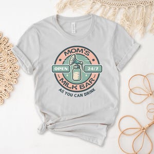 以下が含まれることがあります： ライトグレーのTシャツに、ピンクとグリーンの円形のグラフィックがプリントされています。グラフィックには「Mom's Milk Bar」の文字と、搾乳器のイラスト、そして「Open 24/7」と「All You Can Drink」の文字が書かれています。