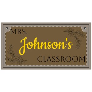 May include: A rectangular sign with a brown background and a gray border. The sign reads "MRS. Johnson's CLASSROOM" in yellow and black text. Decorative leaf branches are in each corner.