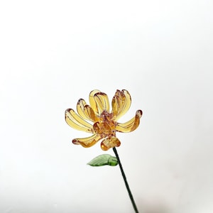 May include: A detailed, amber-hued glass flower with intricate petals and a green leaf. The flower sits on a dark green stem, contrasting against the white backdrop. This decorative item is suitable for interior design and home decor.