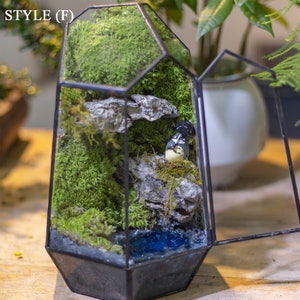 May include: A glass terrarium with a geometric shape, featuring a small figurine, moss, and a blue water feature. The terrarium is open on one side, revealing the interior.