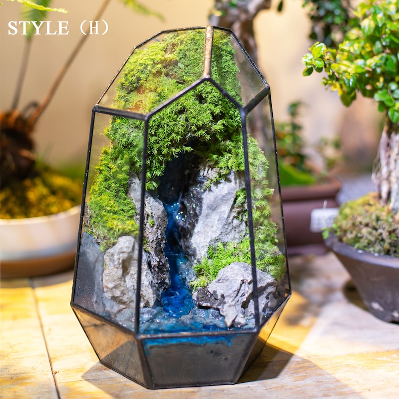 May include: A glass terrarium with a geometric shape, featuring a miniature landscape with moss, rocks, and a blue resin waterfall. The terrarium is labeled "STYLE (H)".