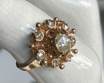 Könnte beinhalten: Ein goldener Ring mit einem großen, runden, facettierten braunen Diamanten, der von kleineren Diamanten umgeben ist. Der Ring hat ein zartes, florales Design.