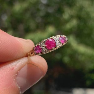 May include: A gold ring featuring three round-cut vibrant pink rubies set in a row, accented by small clear stones embedded in the metal band between the rubies. The band has a textured, decorative finish along the sides of the setting.