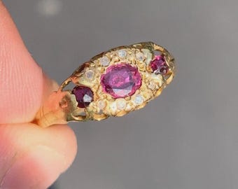 May include: A gold ring featuring a large, oval, magenta-colored gemstone at the center, flanked by two smaller, round, magenta stones. The ring is further adorned with small, clear gemstones. The ring's design is intricate and ornate.