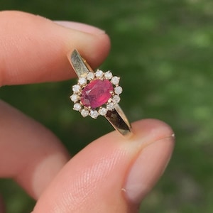 May include: A gold ring featuring a central oval-cut ruby surrounded by a halo of small, round diamonds. The ring's band is a simple, polished gold. The ring is held up against a blurred green background.