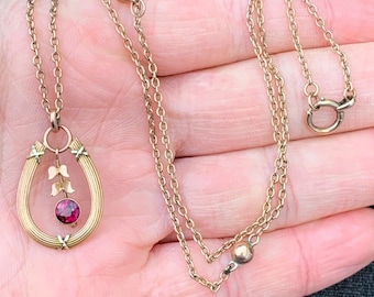 May include: A gold chain necklace with a horseshoe pendant featuring a red gemstone and gold accents.