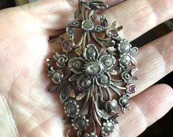 May include: Antique silver-toned pendant necklace featuring a floral bouquet design. The pendant has a central flower with surrounding smaller flowers and accents of purple gemstones. The necklace is held by a delicate chain.