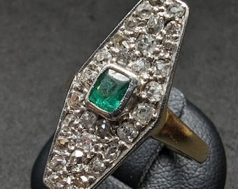 May include: A ring featuring a central rectangular green emerald stone set within a silver-toned elongated hexagonal face. The face is densely encrusted with numerous small, clear, faceted stones surrounding the emerald, mounted on a gold-toned band.