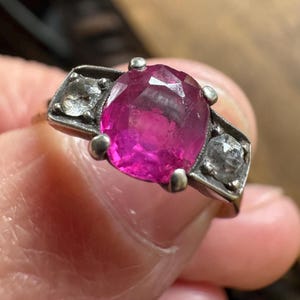 May include: A vintage ring featuring a large, oval-cut ruby gemstone, flanked by two smaller, square-cut diamonds. The ring has a silver-colored setting with a classic, elegant design.