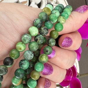 May include: A strand of green gemstone beads, likely jade, with a mottled pattern. The beads are round and smooth, and they are strung on a clear thread. The beads are displayed on a hand with pink nail polish.
