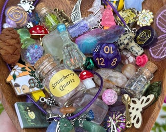 Peut inclure: Une collection de petits charmes et bibelots fantaisistes, dont des bouteilles en verre miniatures, des figurines de champignons, un dé violet et un petit livre. Les articles sont disposés dans un plat en bois en forme de cœur, avec un collier en cordon violet.