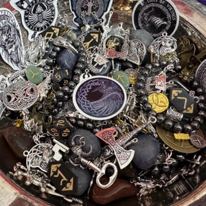 May include: A collection of Viking-themed pendants, charms, and beads in a silver bowl. The assortment includes silver-toned metal pendants with intricate designs, dice, and gemstones. A central oval pendant features a purple and black raven design.