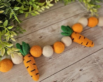 May include: A garland of felt balls in orange and white with two felt carrots with green tops. The garland is on a wooden surface.