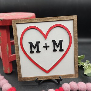 Puede incluir: Un letrero de madera con un corazón rojo y el texto "M+M" en letras negras.