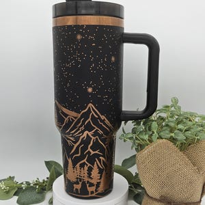 May include: Black travel mug with a copper mountain and forest scene with stars. The mug has a black handle and a black lid.