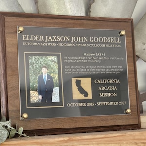 May include: A wooden plaque with a dark brown frame. The plaque features a photo of a person in a suit, text, and a California state outline. The text includes a name, location, and dates.