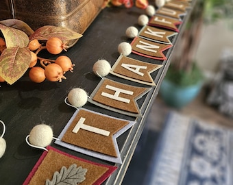May include: A Thanksgiving banner with the word "THANKFUL" spelled out on brown felt pennants with white letters. The banner is decorated with white pom-poms and a leaf design. The banner is displayed on a dark surface with fall-themed decorations.