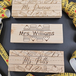 May include: Three wooden teacher nameplates with engraved designs. Each nameplate features a teacher's name: Ms. Dunow, Mrs. Williams, and Ms. Potts. The designs include pencils, apples, scissors, and other school-related icons. Yellow measuring tape surrounds the nameplates.