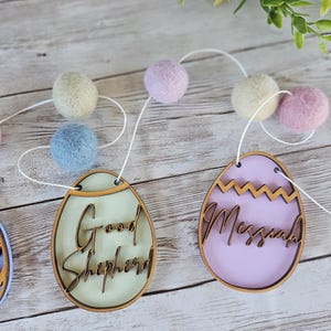 Könnte beinhalten: Eine pastellfarbene Oster-Eiergirlande mit Holzeiern, auf denen "Good Shepherd" und "Messiah" steht. Die Girlande ist mit weißem Garn aufgefädelt und hat pastellfarbene Filzkugeln.