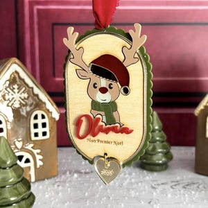 Peut inclure: Une décoration de Noël ovale en bois représentant un renne de dessin animé portant un bonnet de Père Noël rouge et une écharpe verte. Le prénom Olivia est écrit en rouge, avec le texte Mon Premier Noël en dessous et un cœur.