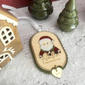 Peut inclure: Une décoration de Noël ovale en bois illustrée d'un Père Noël coloré, avec le prénom "Aubree" en écriture dorée et le texte "Mon Premier Noël". Un petit cœur doré gravé "2016" est suspendu à la base de l'ornement.