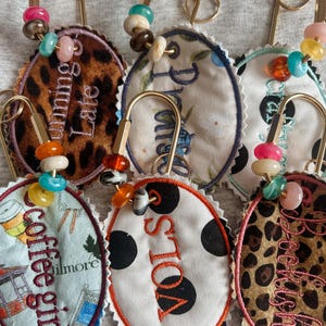 May include: A collection of oval-shaped fabric tags attached to gold-toned safety pins, each adorned with colorful beads. The tags feature various patterns including leopard print, polka dots, and floral designs, with embroidered text such as Running Late, Promise, Coffee Girl, and VOLS.