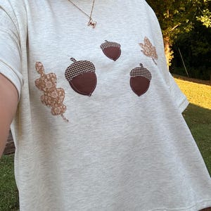 May include: A light heather gray short-sleeve t-shirt featuring an appliqué design of three brown acorns with dark brown and white checkered caps, accompanied by two tan leaves with a white floral pattern stitched onto the front of the fabric.