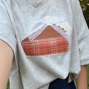 May include: A light gray heathered t-shirt featuring a central appliqué of a slice of pie. The pie slice has a brown top, a white lace trim border, and a crust section made of orange and white plaid fabric. The shirt has short sleeves and a crew neck.
