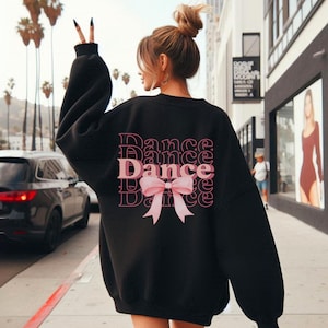 Op de afbeelding: Zwart sweatshirt met een roze strik en het woord "Dance" dat meerdere keren op de achterkant wordt herhaald.
