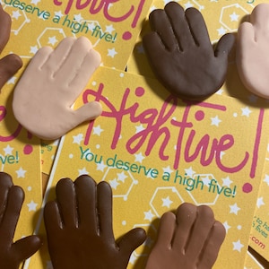 Op de afbeelding: Vier handgemaakte high five handmagneten met een gele achtergrond en witte sterren. De magneten zijn in verschillende huidtinten: lichtbruin, middelbruin, donkerbruin en lichtroze. De tekst op de magneten luidt: "Highfive! Je verdient een high five!"