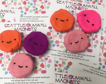 May include: Five colourful, round magnets shaped like a cartoon face with a smile. The magnets are orange, pink, purple, and two shades of pink. The magnets are handmade and feature the text "Seattle Gumwall Magnets".