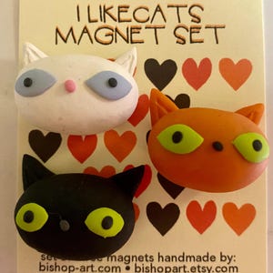 May include: Set of three cat-shaped magnets. The magnets are handmade and feature a white cat with blue eyes, an orange cat with green eyes, and a black cat with green eyes. The magnets are displayed on a background with red and black hearts.