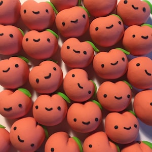 Puede incluir: Una colección de adornos de arcilla de color melocotón con ojos y bocas negros pintados a mano. Cada adorno tiene una expresión facial diferente.