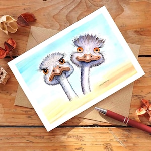 Op de afbeelding: Waterverfillustratie van twee cartoonstruisvogels met expressieve ogen op een witte kaart. De kaart ligt op een bruine envelop, met een rode pen en gedroogde bloemen op een houten oppervlak.