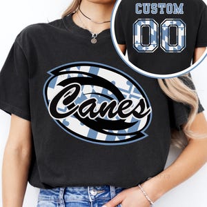 Könnte beinhalten: Schwarzes T-Shirt mit einem großen ovalen Design mit dem Wort "Canes" in weißer Schrift, umrandet in Schwarz. Das Oval hat ein blau-weißes Muster. Auf der Rückseite des Shirts steht "CUSTOM" über der Nummer "00" in einem blau-weißen Muster.