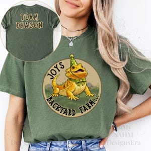 May include: An olive green t-shirt featuring a circular graphic of a yellow bearded dragon wearing a green party hat and bow tie. The text reads JOYS BACKYARD FARM LLC. A circular inset shows the back of the shirt with the text TEAM DRAGON.
