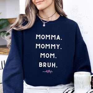以下が含まれることがあります： ネイビーブルーのスウェットシャツに、白い文字で「Momma. Mommy. Mom.  Bruh.」と書いてあり、その下にピンクのハートと「mom life」の文字があります。
