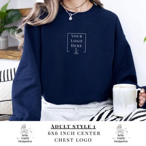 May include: A navy blue sweatshirt featuring a white square with the text "YOUR LOGO HERE". The text "ADULT STYLE 1 6X6 INCH CENTER CHEST LOGO" is printed below. The sweatshirt is a solid colour.